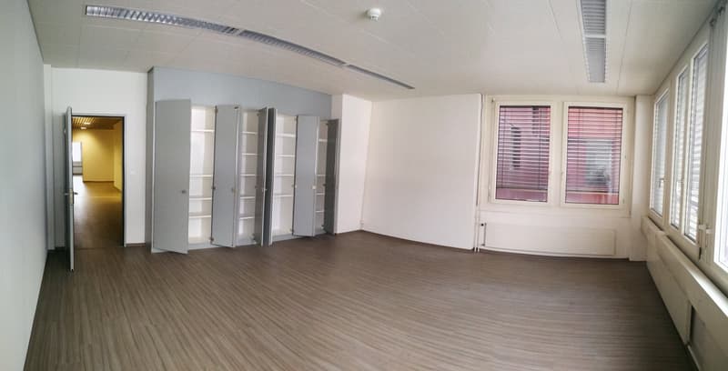 Large open office space with white walls, gray laminate flooring, and multiple storage cabinets along the walls. The room has large windows providing natural light and a doorway leading to another room.