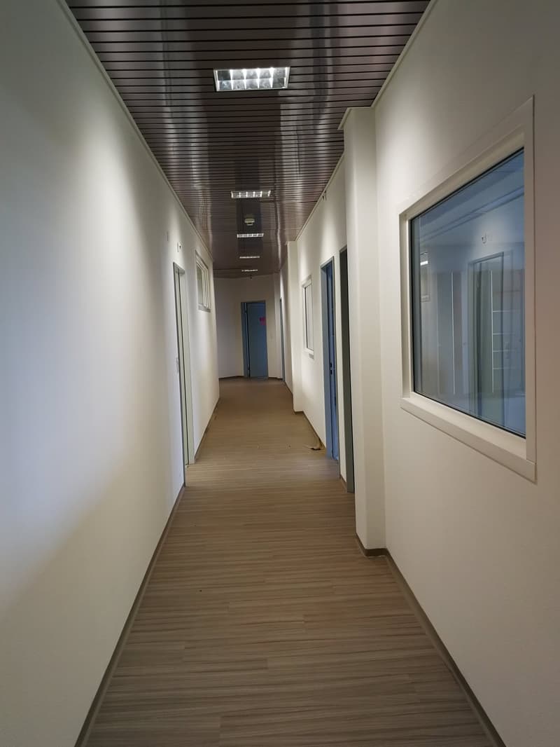 The image shows a long, narrow hallway with a wooden floor and white walls. The ceiling is made of dark wooden panels, and there are recessed lighting fixtures installed along the ceiling. The hallway has several doors leading to other rooms or offices, a