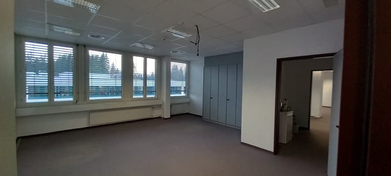 Large open office space with multiple windows, grey walls, suspended ceiling, and storage lockers along one wall.