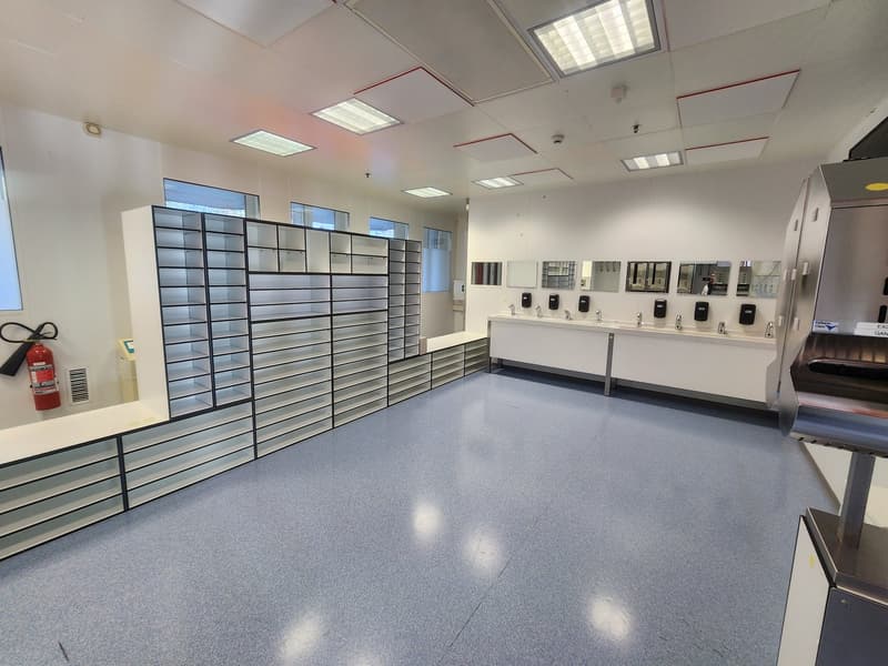 Empty room with white walls and gray flooring, shelves with compartments, fire extinguisher, sinks with faucets and soap dispensers, mirror.