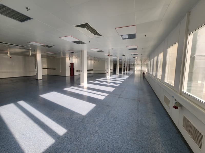 A long hallway with large windows on both sides, gray floor, white ceiling, and columns.