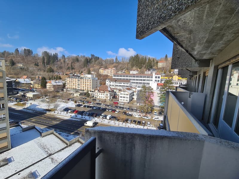 A balcony with a view of a town with many buildings, a road, and a parking lot.