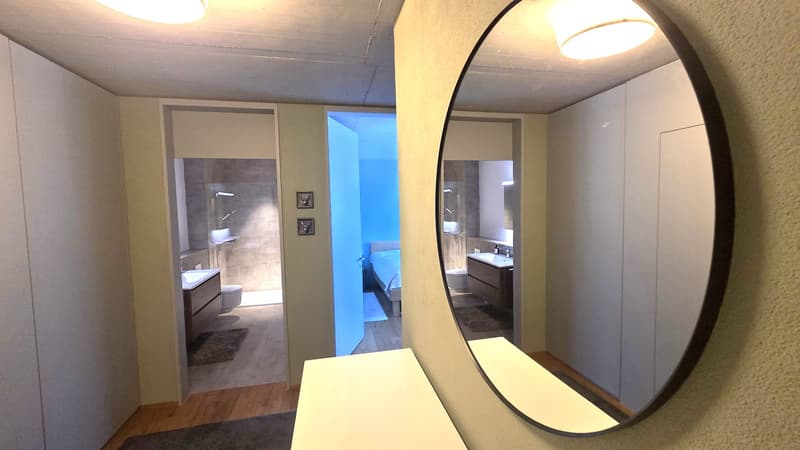 A hallway with a mirror, a wooden floor, and a white wall. The mirror reflects the bathroom and bedroom. The bathroom has a sink, toilet, and shower. The bedroom has a bed, a cabinet, and a mirror. The hallway has a rug and a light on the ceiling.