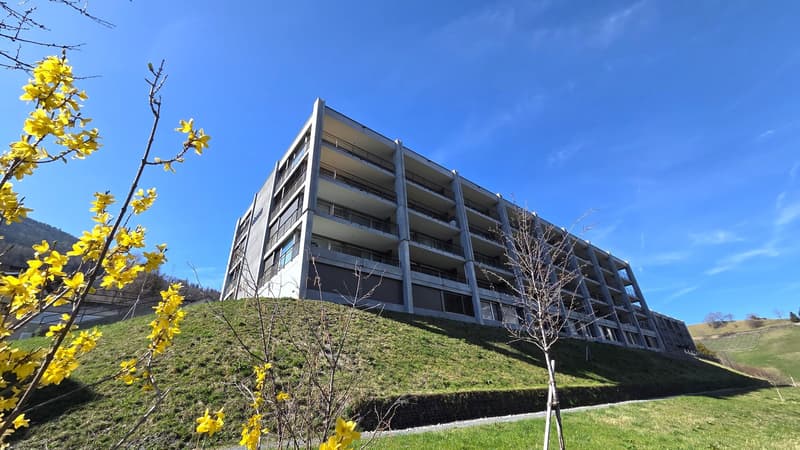 Modern apartment building, multiple floors, balconies, concrete and glass exterior, set on a hill