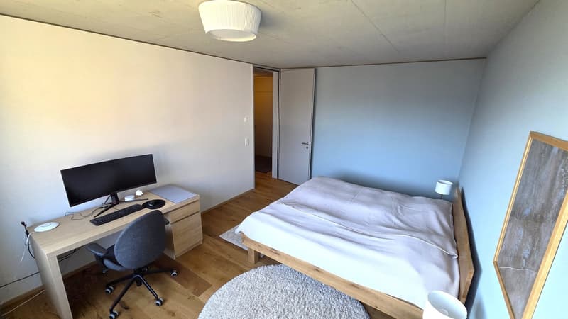 A bedroom with a bed, desk, chair, monitor, keyboard, mouse, lamp, rug, and a door leading to another room.