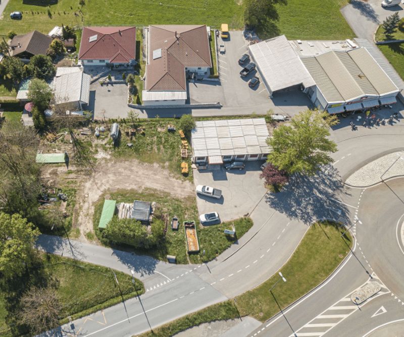 The image shows an aerial view of a residential area with several houses, a road, and a parking area. The houses have red tile roofs and the surrounding area is covered in green grass and trees.