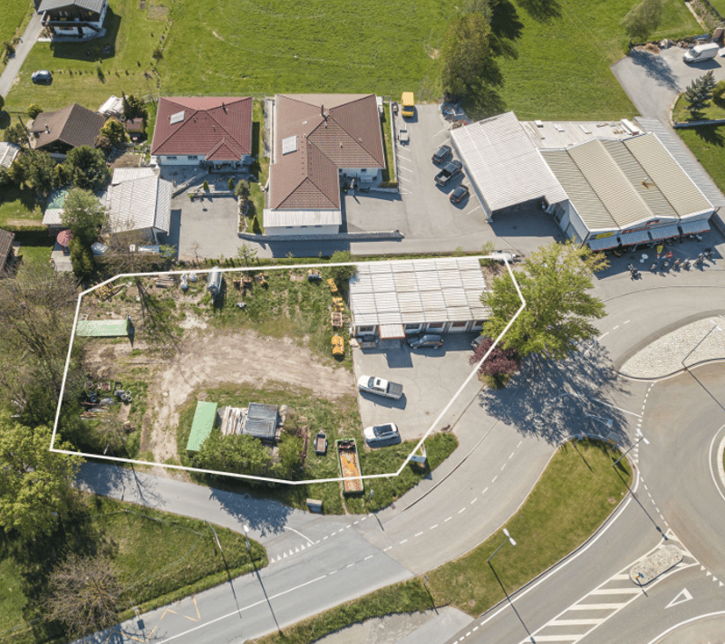 The image shows an aerial view of a residential area with several houses, a parking lot, and a road. The houses have red tile roofs and appear to be two-story buildings. There is a parking lot with several vehicles parked in it, and a road running through