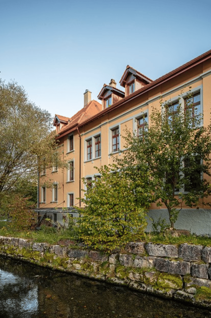 3 story yellow building, multiple windows, stone wall, river in front