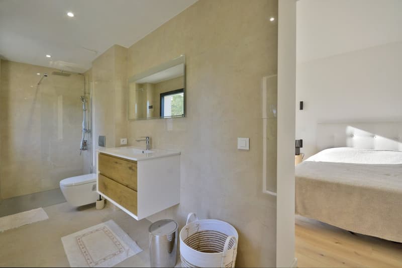 Bathroom with shower, toilet, sink, and wooden cabinet. Adjacent to a bedroom with a bed and a nightstand.