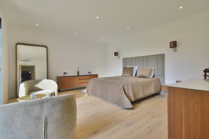 Bedroom with a large bed, wooden floor, white walls, a mirror, a wooden cabinet, and two chairs.