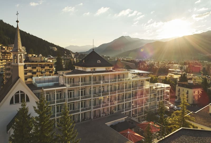 Multi-story building with balconies, surrounded by mountains and trees, with a church-like structure in the foreground and a scenic mountain landscape in the background.