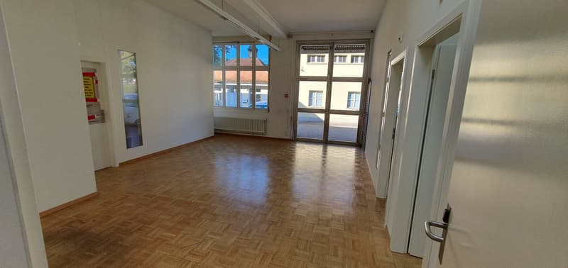 Empty room with wooden floor, white walls, glass doors, windows, radiator, and a hallway leading to other rooms.