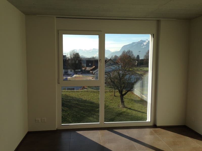 Spacious room with large windows overlooking a scenic outdoor landscape. The room has a tiled floor and neutral-colored walls, providing a clean and minimalist aesthetic. The view through the windows showcases a grassy area with a tree and distant mountai