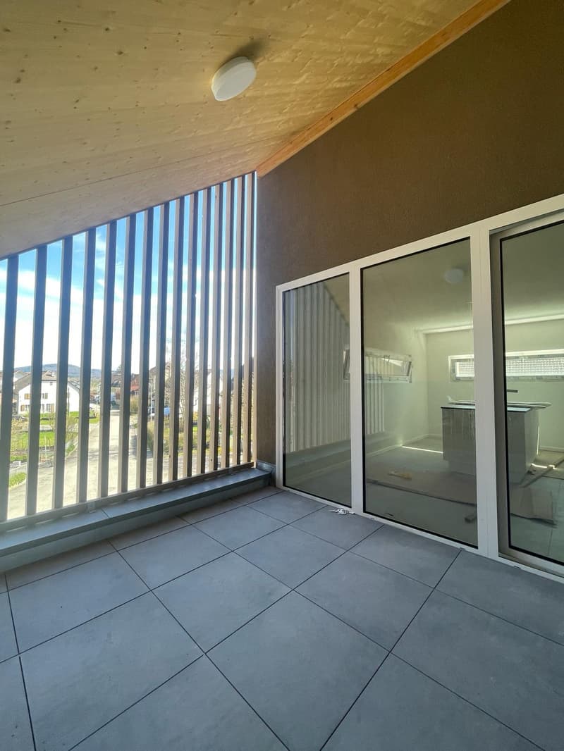 This image shows a room with a balcony or terrace. The room has a wooden ceiling with exposed beams, and the floor is covered with gray tiles. There are large windows along one wall, providing a view of the outdoors. The room appears to be a living or din