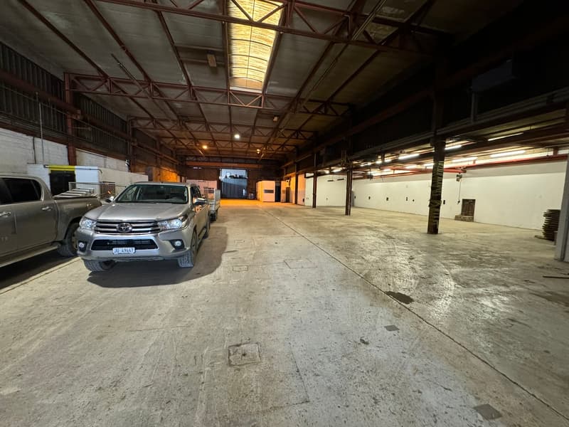 Warehouse with concrete floor, metal beams, open space, lights on the ceiling, parking space for vehicles, and vehicles parked.