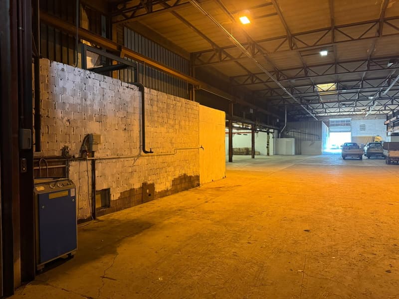 Large empty warehouse with metal frame ceiling, yellow walls, electrical equipments on left, open entrance with cars parked