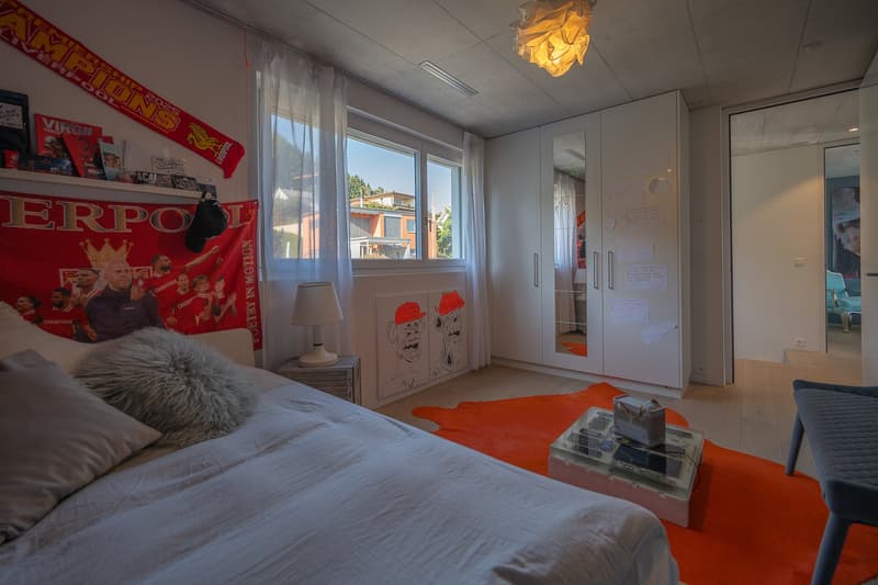 Bedroom with bed, lamp, rug, and wardrobe. Wall decorations include a red banner and a shelf with books and a hat.