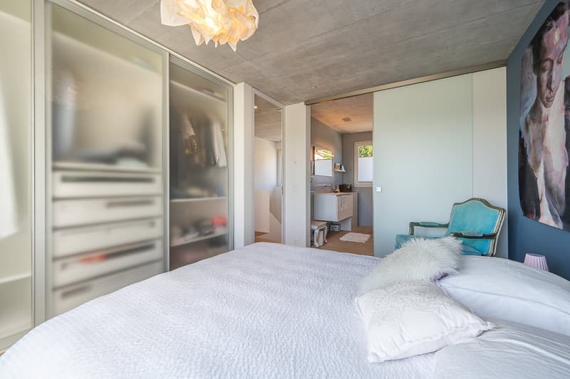 large bed, white linens, white pillows, glass closets, white walls, white ceiling, chandelier, blue chair, painting