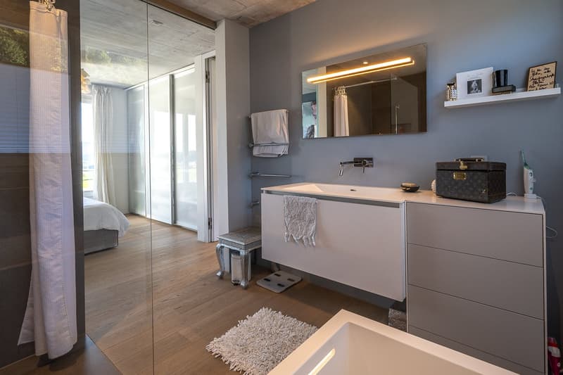 modern bathroom, white sink, large mirror, glass shower, wooden floor, white rug, white cabinets, shelves with personal items