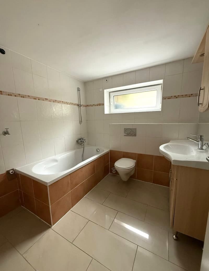 Spacious bathroom with white and brown tile walls, a bathtub, a sink, and a toilet. The bathroom has a window for natural light.