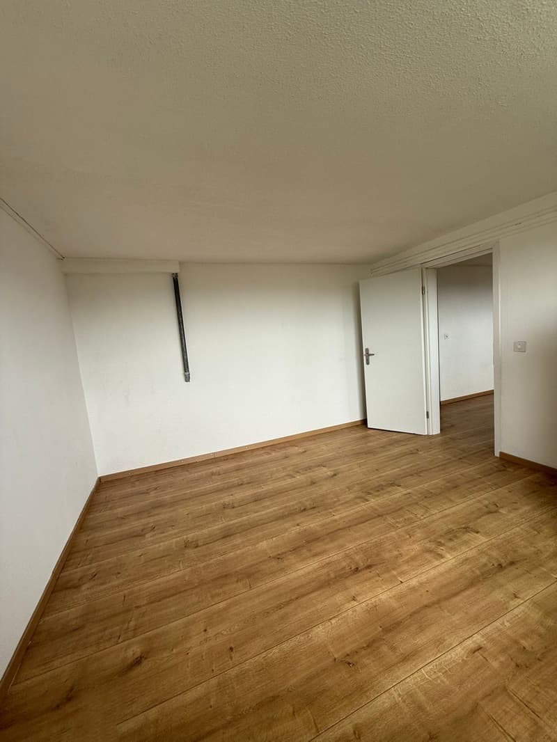 Empty room with white walls and hardwood flooring. The room has a door visible and appears to be a living or bedroom space.