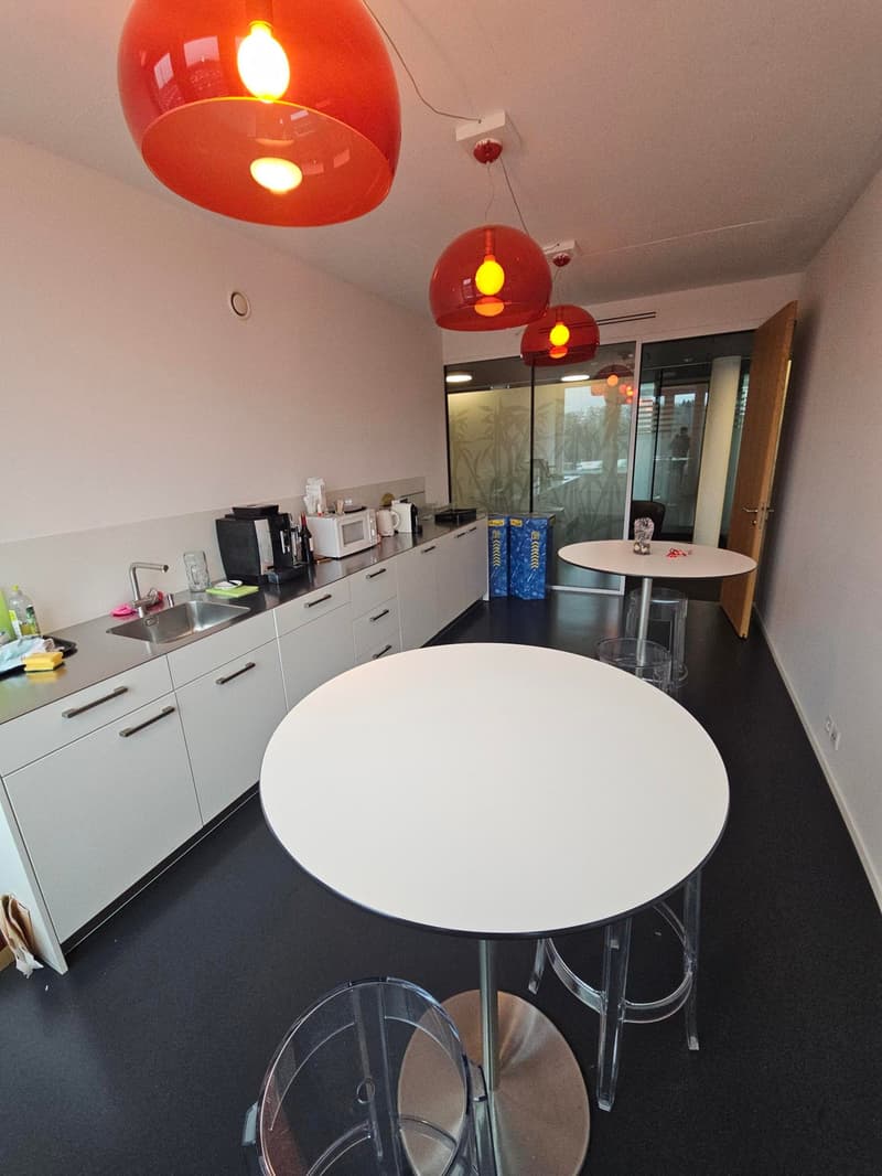 White kitchen with a central island, black countertops, stainless steel sink, red pendant lights, coffee machine, microwave, trash bins, and clear bar stools.