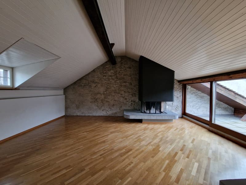 Spacious attic living room with exposed stone wall, hardwood flooring, and a fireplace. Large windows provide natural light and a view of the exterior.