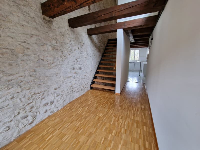 The image shows the interior of a room with a stone wall and exposed wooden beams on the ceiling. The floor is made of hardwood planks. There is a staircase leading up to another level. The room appears to be part of a larger living space or apartment.