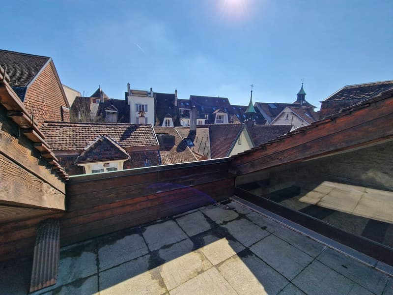 The image shows a view of a historic European city, with a cluster of traditional buildings with red tiled roofs and ornate architectural details. The buildings appear to be situated on a hillside, with a blue sky and sun visible overhead. The scene has a