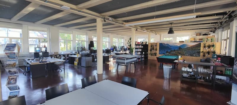 Large open-concept office space with high ceilings, exposed beams, and large windows. The space includes desks, computers, bookshelves, and a pool table. There is also a fireplace visible in the room.