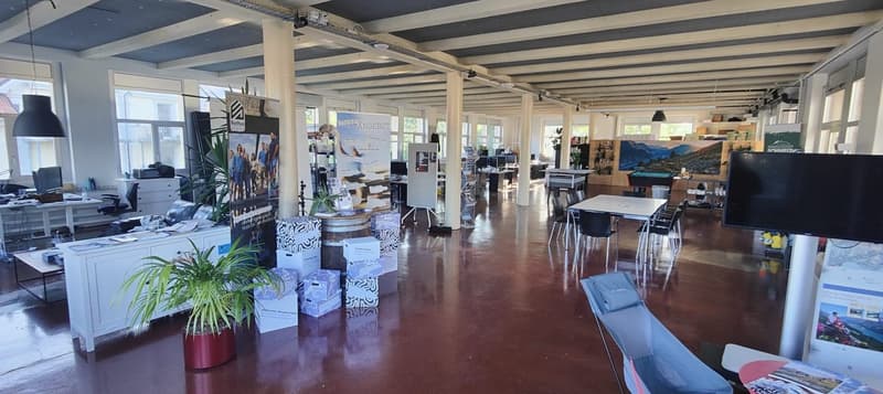 Large open-plan office space with high ceilings, exposed beams, and concrete floors. The space is filled with desks, chairs, and various office equipment and supplies. There are also some plants and decorative elements throughout the space.