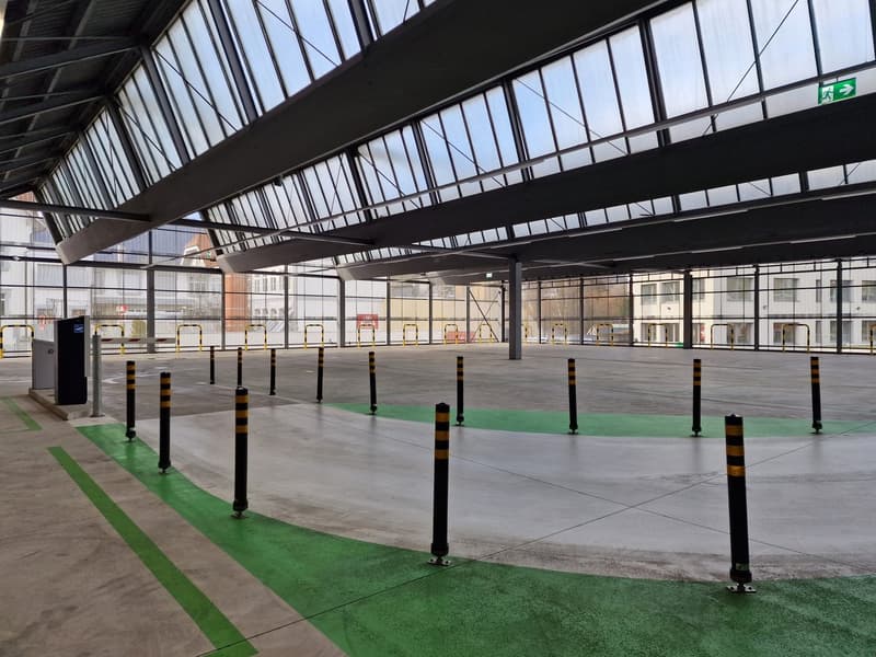 Large industrial warehouse with high ceilings, glass walls, and concrete flooring. The space has designated parking areas marked with green lines and black and yellow bollards.