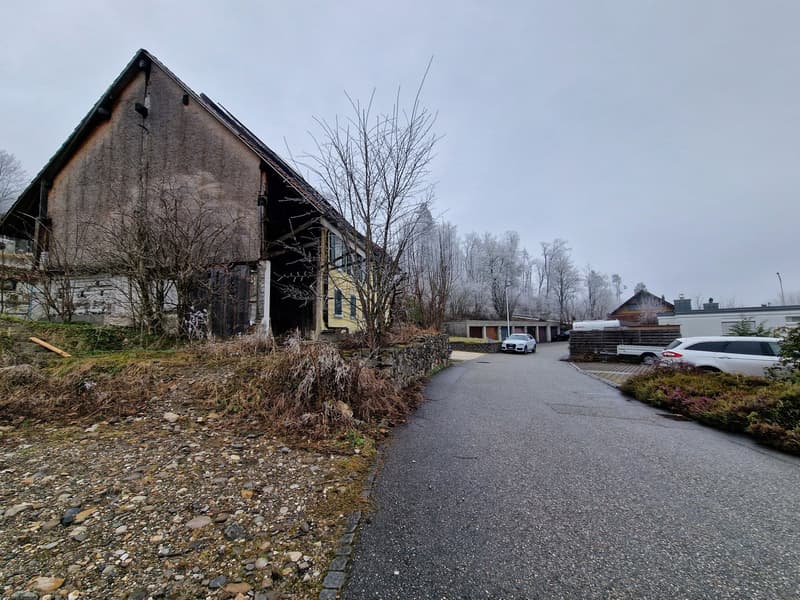 An old, dilapidated barn or warehouse structure with a slanted roof and weathered exterior. The building is surrounded by bare trees and a paved driveway or parking area, with other smaller structures visible in the background.