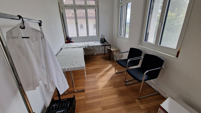 White walls, wooden floor, desks with white cloths, two black chairs, three windows, a hanging rack with a white shirt, boxes, and a monitor on a table.