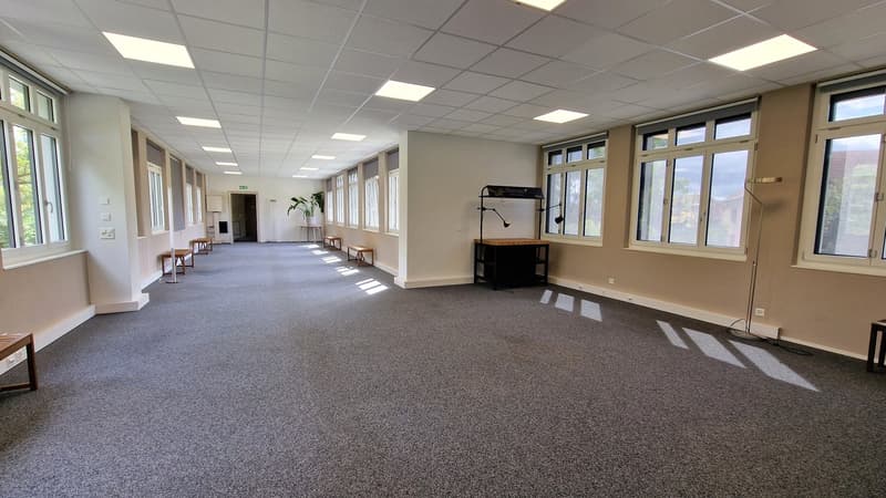 Empty room with wooden floors, bright ceiling lights, wooden benches, and large windows.