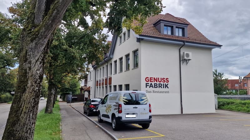 white facade, brown roof, several windows, two cars parked, trees in the front, text sign