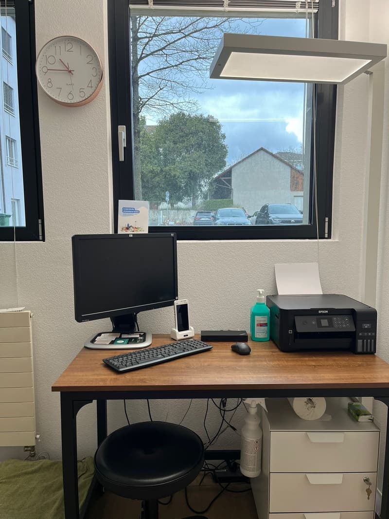 A desk with a computer monitor, keyboard, mouse, and printer. A clock is on the wall, and a window is behind the desk. A radiator is on the left side, and a stool is in front of the desk. A bottle and a piece of paper are on the desk.