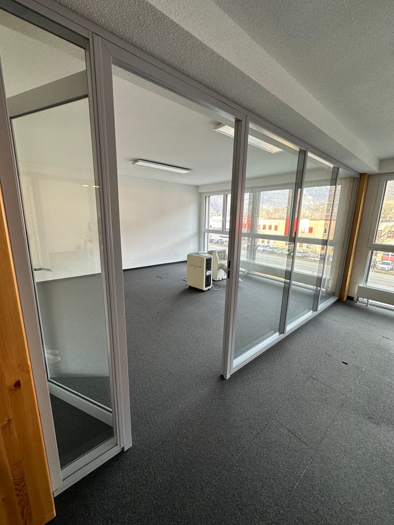 The image shows a large open office space with floor-to-ceiling windows along one wall, providing ample natural light. The room has a gray carpeted floor and white walls. There is a portable air conditioning unit visible in the corner of the room.