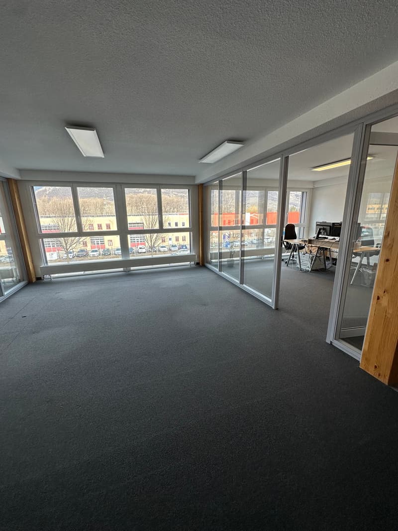 Large open office space with floor-to-ceiling windows providing a view of the surrounding buildings and landscape. The room has gray carpeted flooring and recessed lighting fixtures on the ceiling. There is a desk and chair visible in the corner of the ro