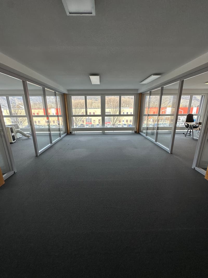 Large open office space with floor-to-ceiling windows, grey carpeted floor, and recessed lighting in the ceiling.