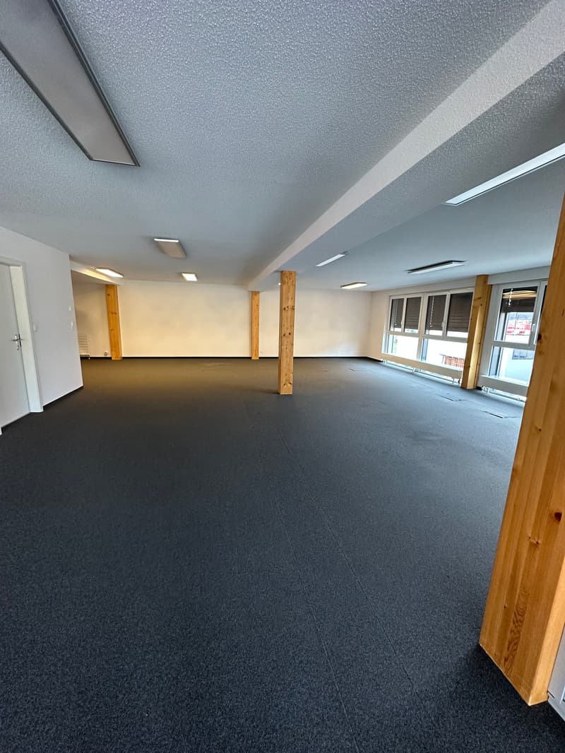 Large open office space with gray textured ceiling, white walls, and dark gray carpeted flooring. The space has several wooden support columns and recessed lighting fixtures.