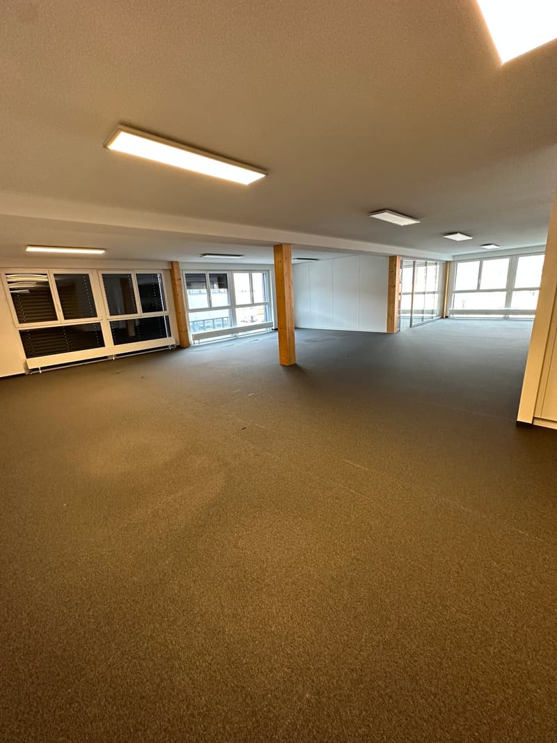 Large open office space with carpeted flooring, white walls, and recessed lighting. The space has several windows providing natural light and wooden support columns.
