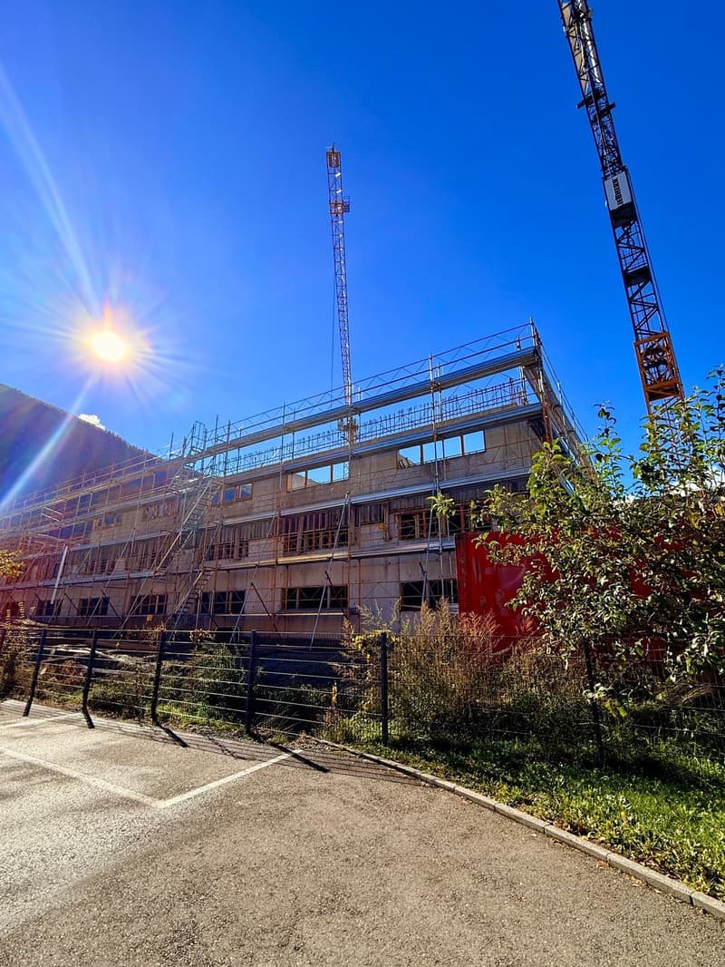 A building under construction, several stories, windows, scaffolding, cranes, a large view of the sun and mountains, an empty parking area.