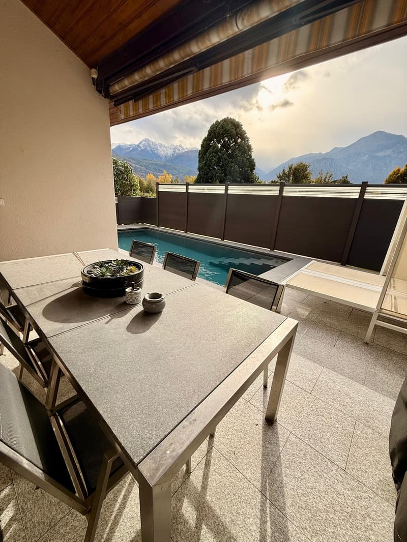 Terrace with a dining table and chairs, surrounded by a pool and fence, overlooking mountains
