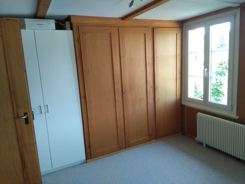 An empty room with wooden wardrobes and a white wardrobe against the wall, a window with a white frame and a white radiator below it.