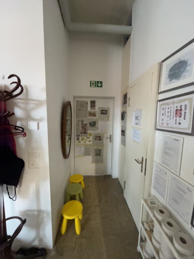 Corridor with white walls, yellow and green stools, mirror, bulletin board, and papers. Exit sign is on the door. A roll of toilet paper is on a shelf on the right.
