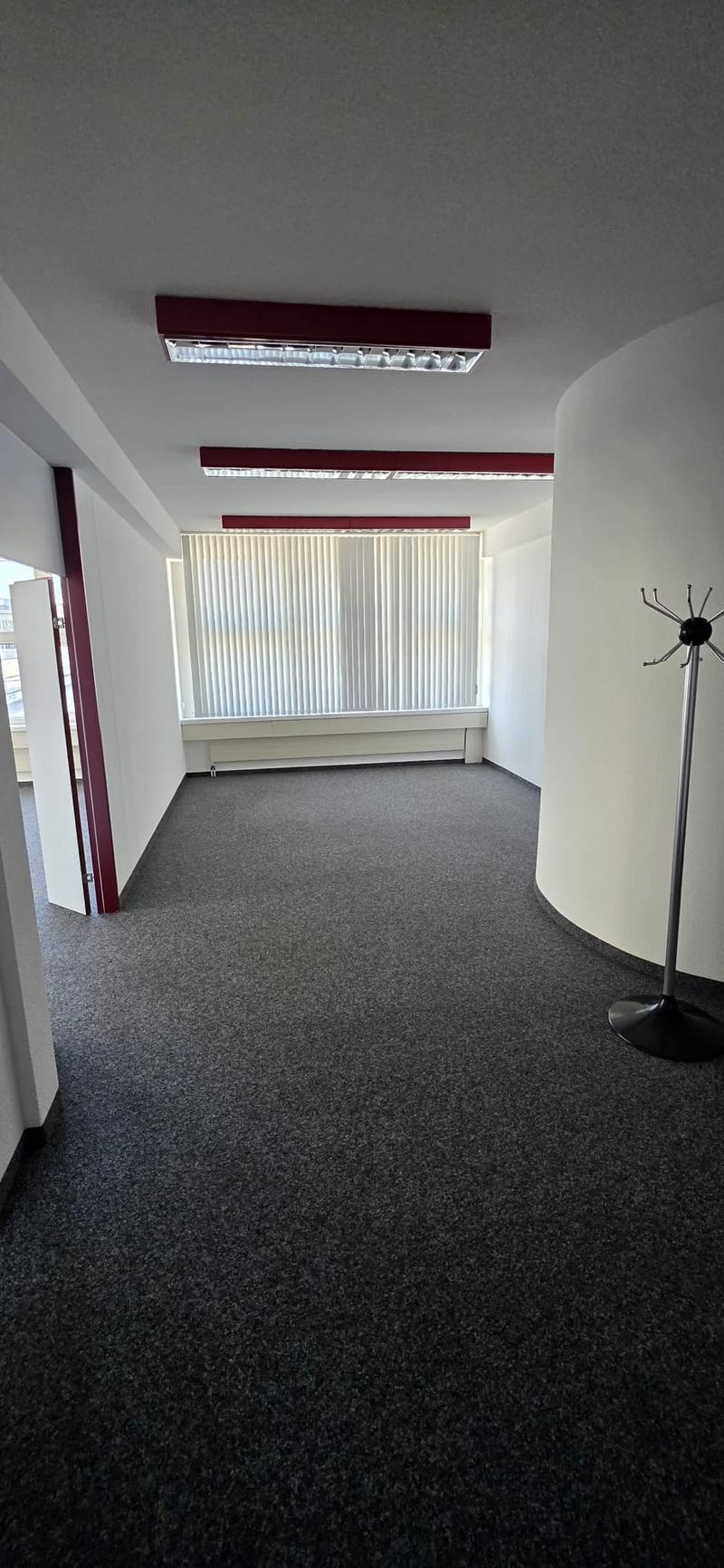 Large empty room, white walls, gray floor, window with white blinds, track lighting, coat rack