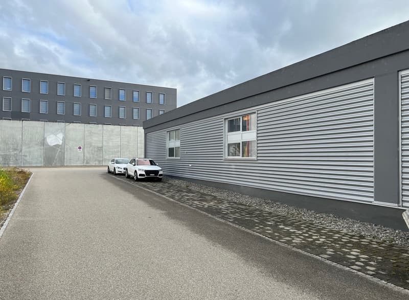 Two grey buildings, concrete parking lot, 2 white cars parked, gravel pathway, cloudy sky