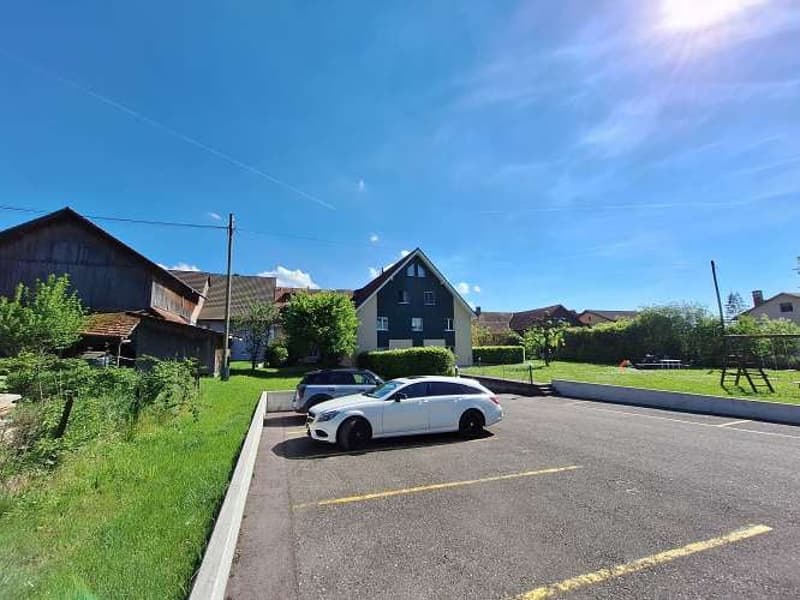A parking lot with 2 parked cars, surrounded by a grass field and shrubs, behind the parking lot there are residential buildings