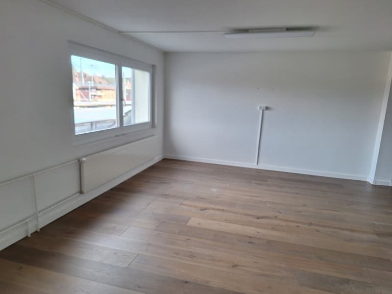 Empty room with large window, wooden floor, wall-mounted radiators, and a ceiling light.